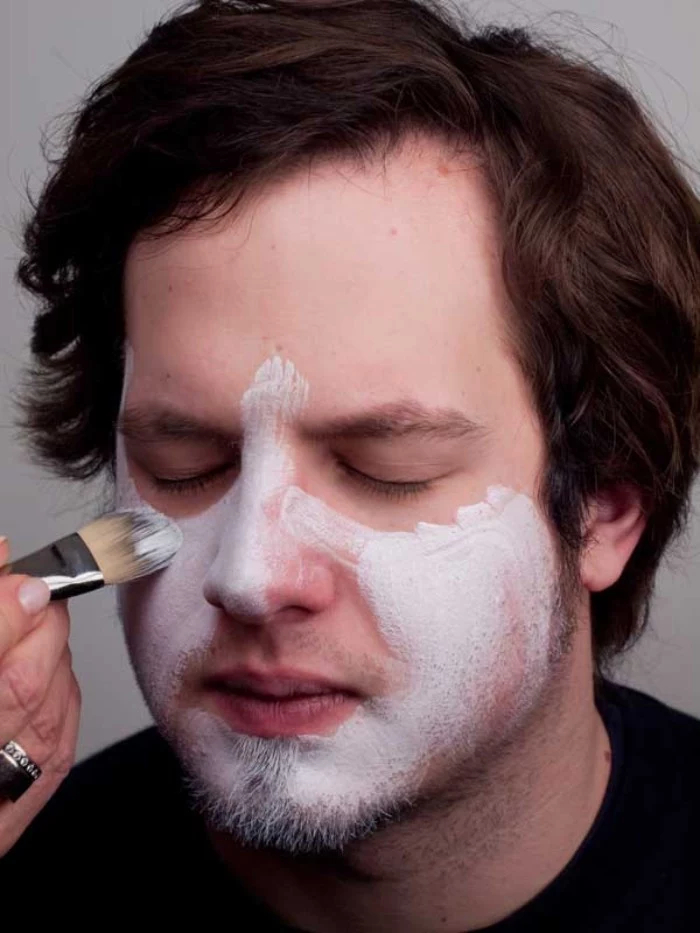 clown face paint, recreating the look of the joker from batman, hand applying white paint, on the face of a young man, with wavy brunette hair