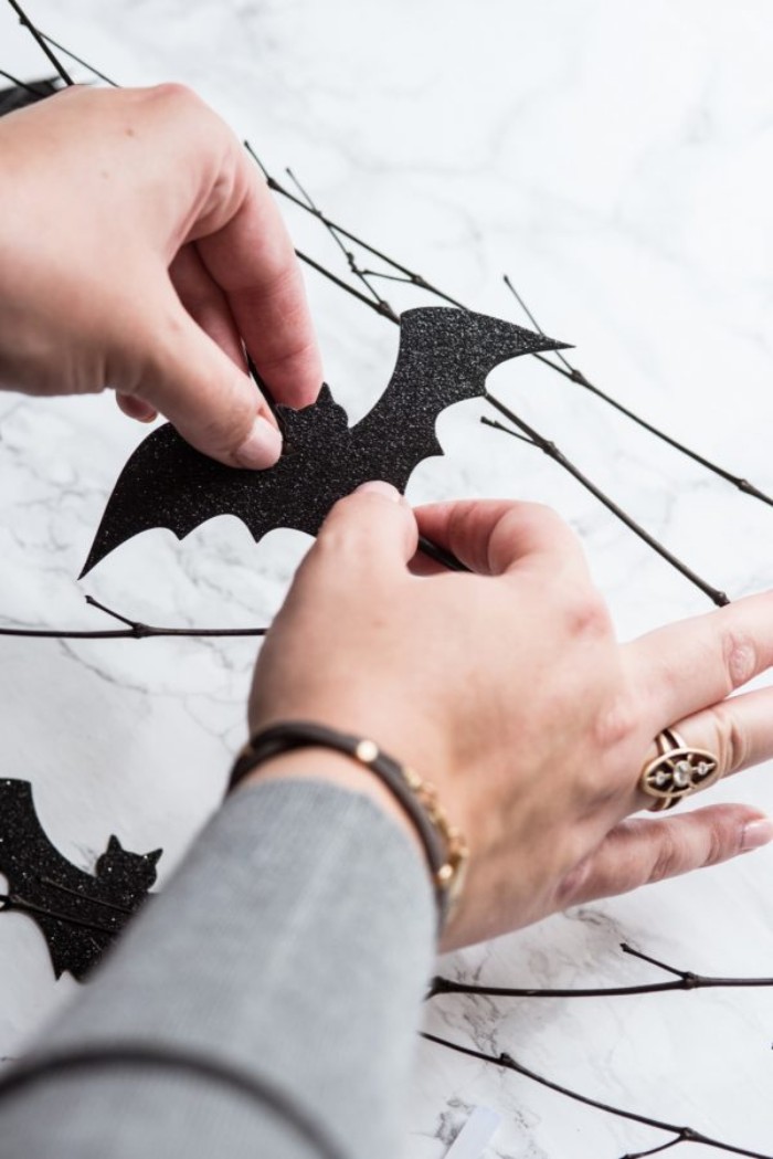 gluing a black, bat-shaped card cutout, onto a dark brown, thin dried branch, placed on a light marble surface