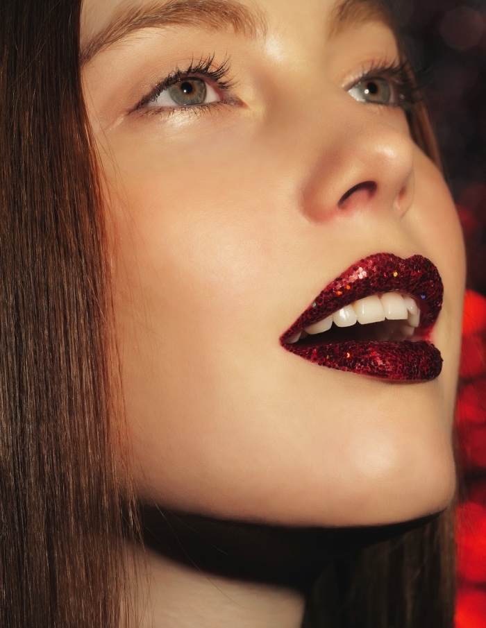 flakes of red glitter, worn over red lipstick, christmas makeup ideas, on a young and slim, brunette woman with blue eyes