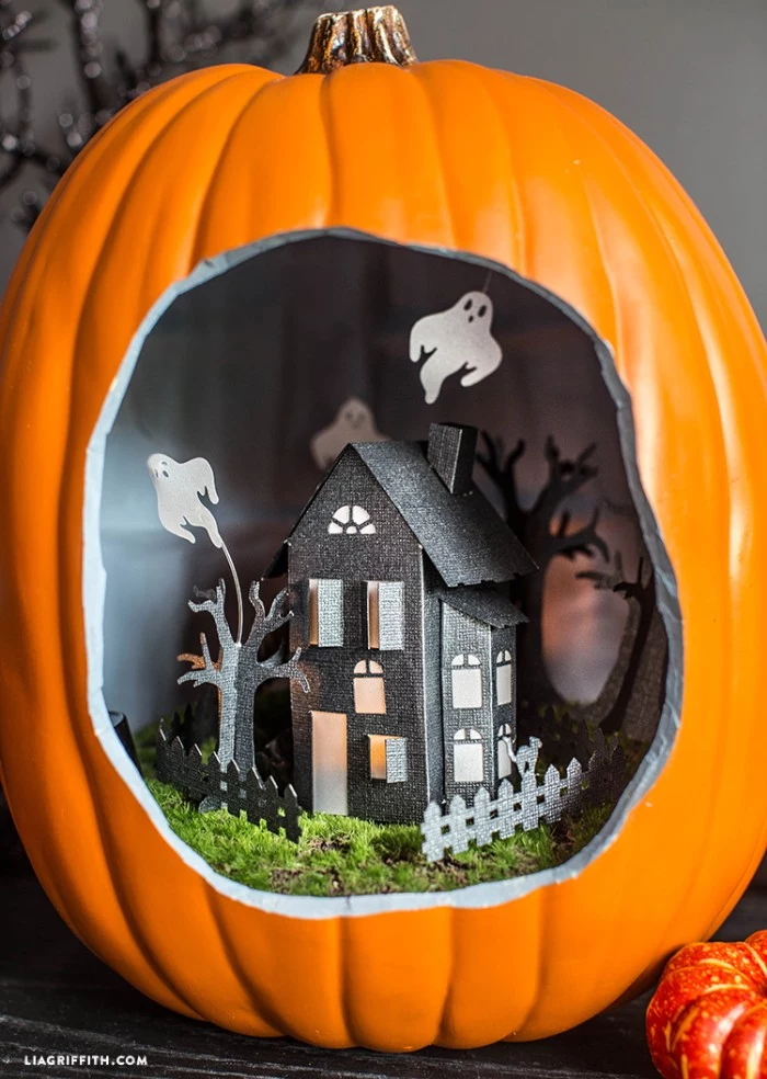 decorative ornament shaped like a pumpkin, with a large hole, revealing a small paper house inside, decorated with three paper ghosts, halloween decorations