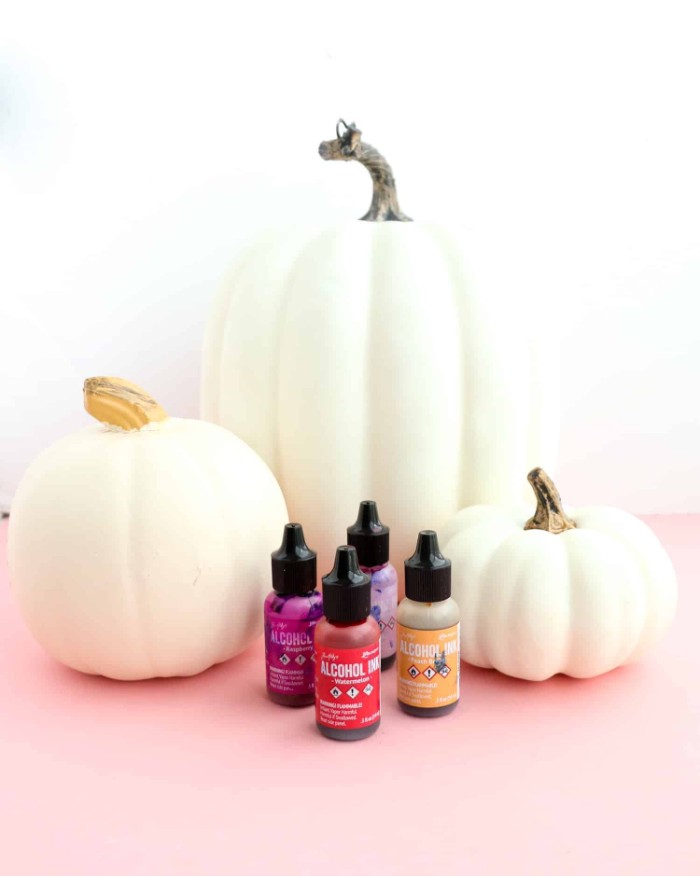 halloween pumpkin decorations, three white fake pumpkins, placed on a pale pink surface, near four small bottles of paint, in differen colors