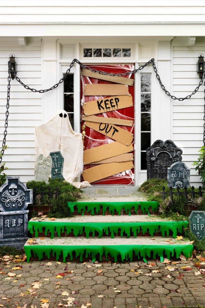 red door boarded with pieces of cardboard, with the words keep out, written in black runny paint, scary outdoor halloween decorations, stairs covered with green stickers, looking like slime, chains and fake gravestones