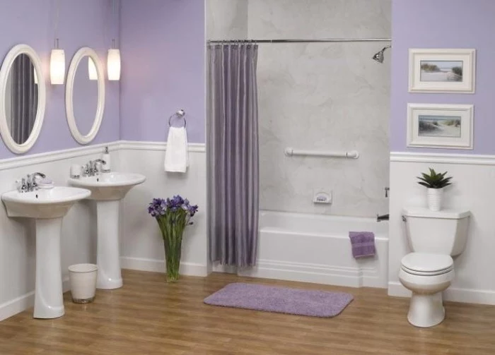 a set of identical white sinks, and two oval mirrors in white frames, in a room with pale lavender walls, and white wood paneling, bathtub and toilet