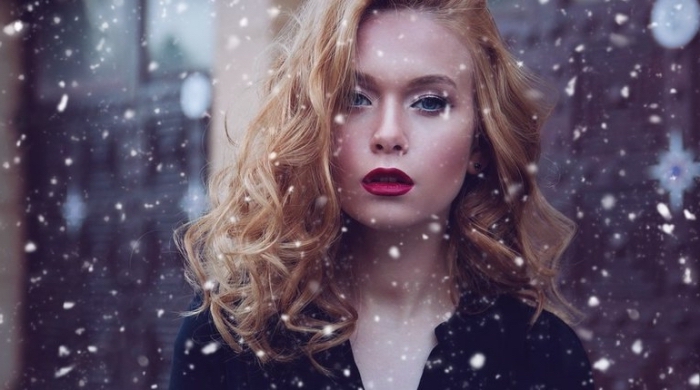 snow falling aroud a young woman, with curly blonde hair, wearing dark red lipstick, and a black top