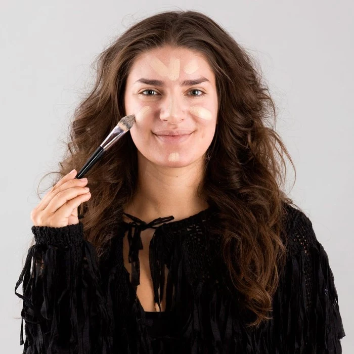 smiling brunette woman, with long and wavy brunette hair, applying foundation on her face, using a brush, halloween face paint how to