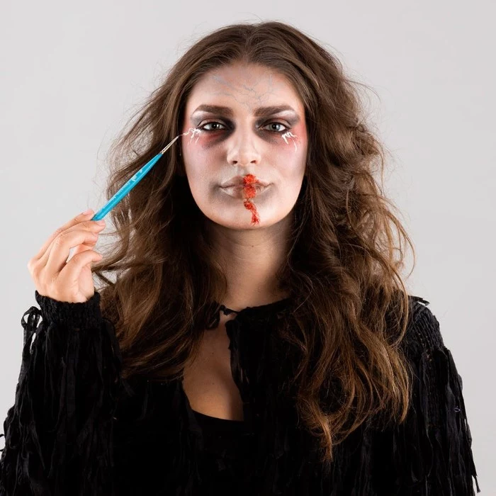 teal brush used to add special effects makeup wax, near the corners of the eyes of a young woman, with zombie face paint, and brunette wavy hair
