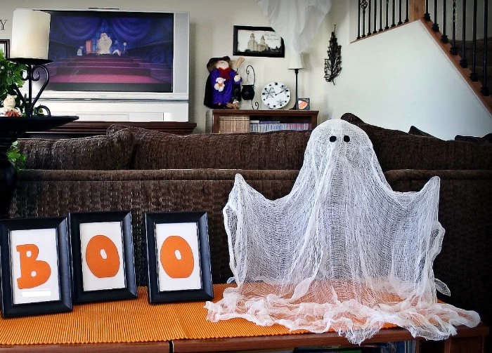 the letters B O O, written in orange, in three black frames, on a table near a ghost decoration, made from gauze and glue, scary halloween decorations
