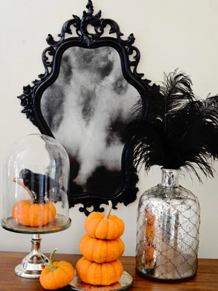 baroque frame in black, on a mirror, decorated with an image of grey smoke, haunted house decorations, several small pumpkins, a silver bottle with black feathers, a glass dish with a pumpkin, and a crow statuette