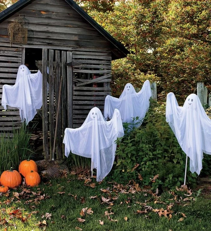 ghosts made of sheer fabric, stuck on wooden poles, with arm-like parts, and simple hand-drawnfaces, in a garden with a broken, old wooden shed, scary outdoor halloween decorations