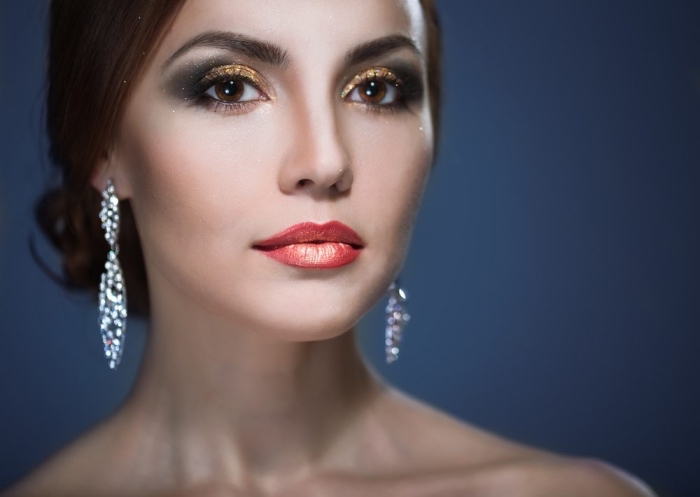 christmas eye makeup, in gold with smokey effect, worn by a pale woman, with brunette hair, styled in a bun, glossy orange lipstick, and sparkling silver earrings