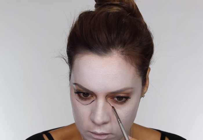 brunette woman with face covered in white paint, using a small brush, to outline her eyes, skeleton face paint, steb by step