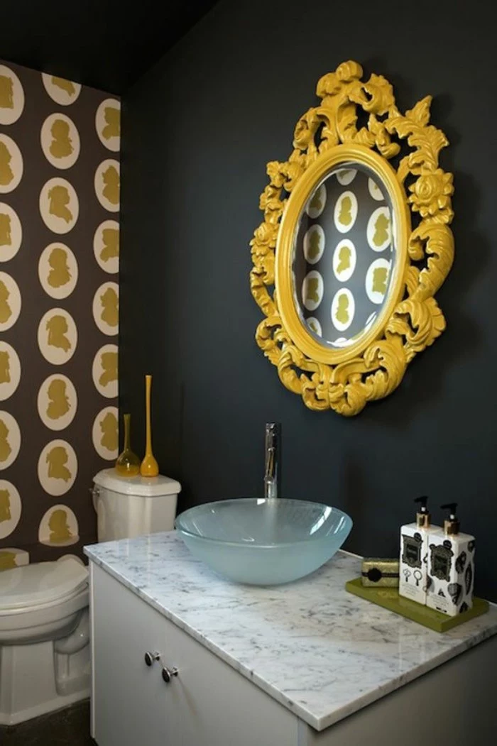 baroque frame in yellow, around an oval mirror, mounted on a black wall, small bathroom paint colors, over a white cupboard, with a marble countertop, and a small sink