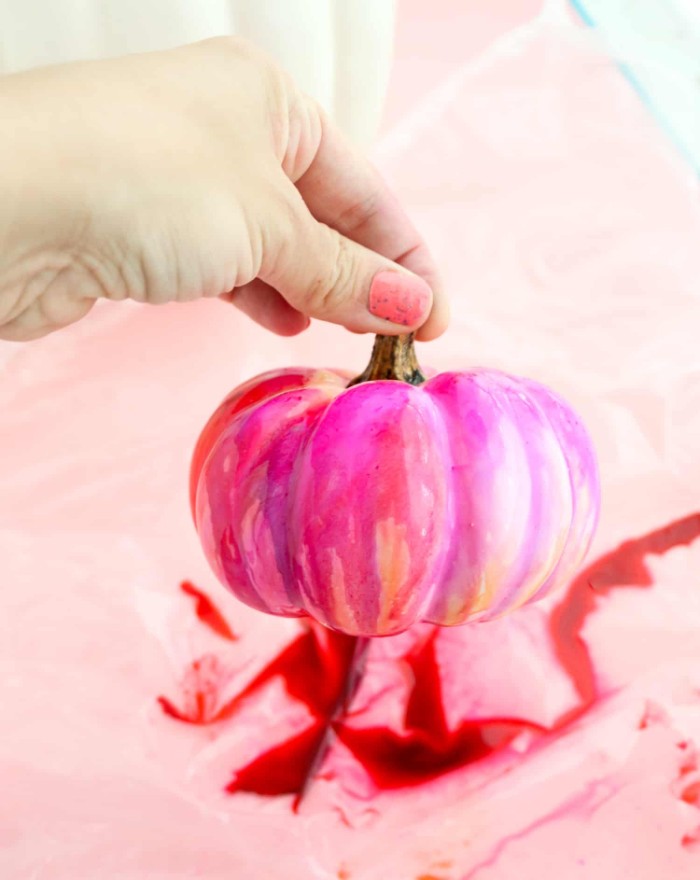 hand with pink nail polish, holding a small fake pumpkin, painted in pink, red and orange, halloween pumpkin decorations, over a piece of plastic with red paint