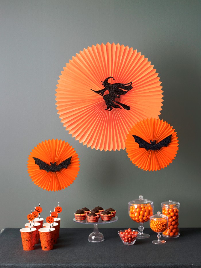 halloween party decoration ideas, three round orange paper ornaments, decorated with black cutouts, shaped like a witch flying on a broomstick, and two bats, hanging over a table, with snacks and drinks