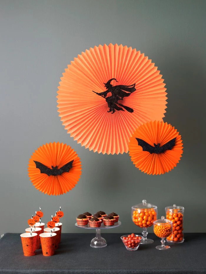 halloween party decoration ideas, three round orange paper ornaments, decorated with black cutouts, shaped like a witch flying on a broomstick, and two bats, hanging over a table, with snacks and drinks