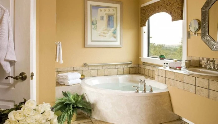 corner bathtub in cream, inside a bathroom with pale, pastel orange walls, decorated with beige tiles, white roses and a potted fern