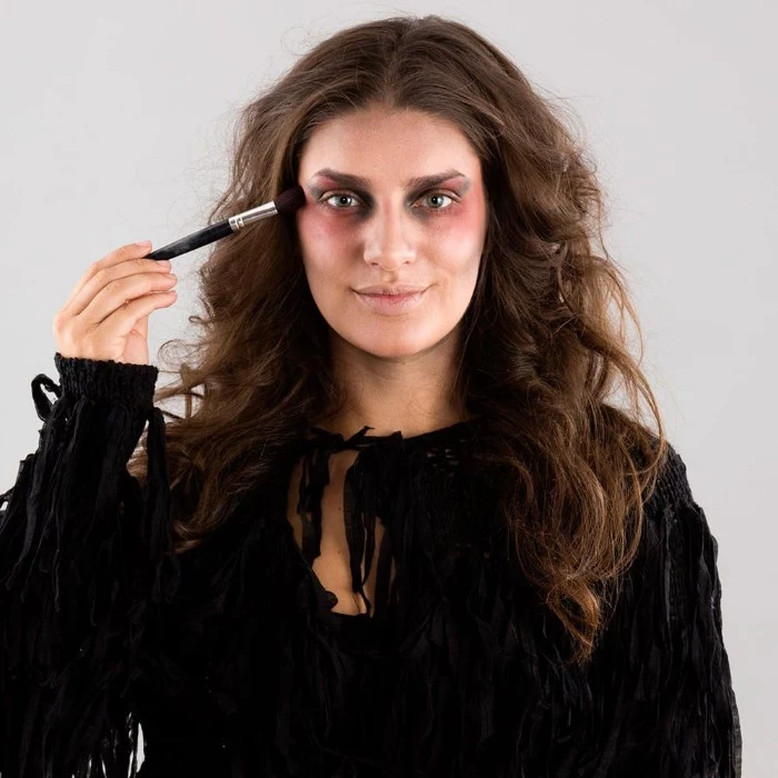 zombie face paint, tutorial with photos, smiling brunette woman, dressed in black, putting pink blush, around her eyes, using a brush