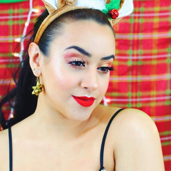 rose gold eye shadow, worn with fake eye lashes, bright red lipstick, and shimmering blush, christmas eye makeup, on a smiling woman, with dark hair, anc christmas-themed accessories
