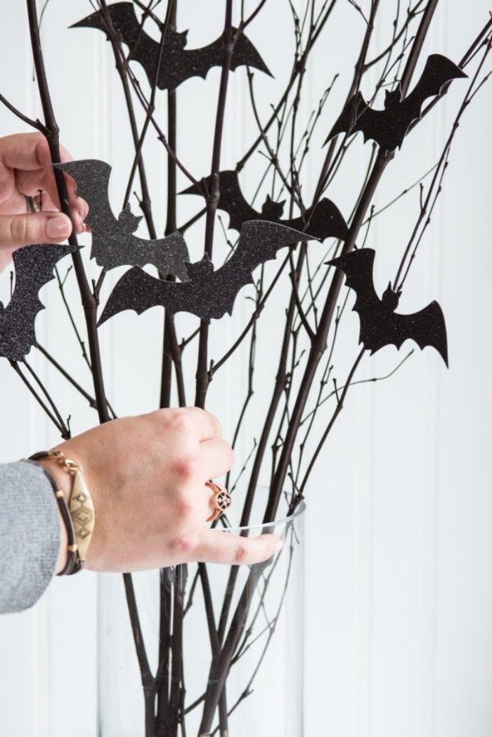 putting the ready halloween decoration, consisting of thin dried branches, with small glittering black bat shapes, in a clear glass vase