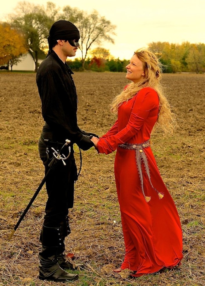 buttercup and wesley, costumes inspired by the film the princess bride, man dressed in black, with a mask on his face, holding the hands, of a woman with long curly blonde hair, wearing a red gown