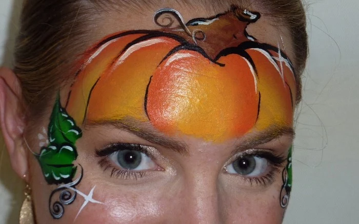 a different kind of witch face paint, pumpkin painted in orange and black, brown and white, with little green leaves, on the forehead of a young woman