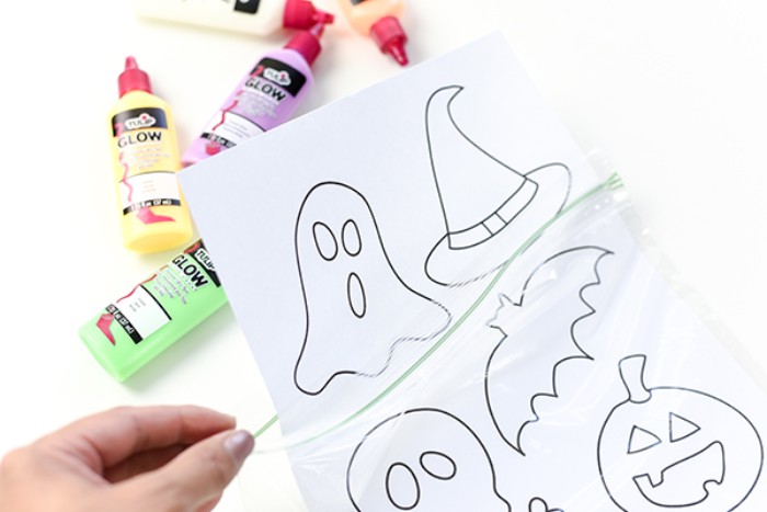 sheet of white paper, with simple drawings of halloween related objects, placed in a ziploc bag, held by a hand, with pale pink nail polish, halloween decorations diy, tubs of paint in the background