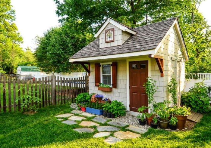 cottage style she shed, tiny house with a brown door, and a window with wooden shutters, inside a garden, with various potted plants