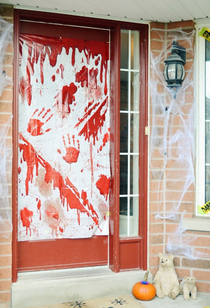 bloody arm prints and splatters, painted on a sheet of white plastic, stuck to a door, scary outdoor halloween decorations, fake cobwebs and a pumpkin nearby