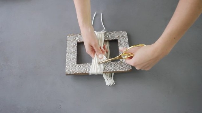 brown picture frame, with white details, and yarn wrapped around it, room makeover with a diy chandelier, hands with scissors, cutting the yarn free from the frame