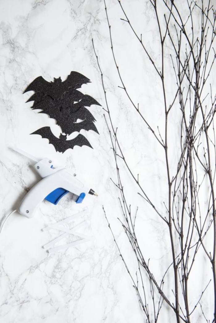 glue gun and several black, bat-shaped card cutouts in black, on a marble surface, near a bunch of thin, dried tree branches