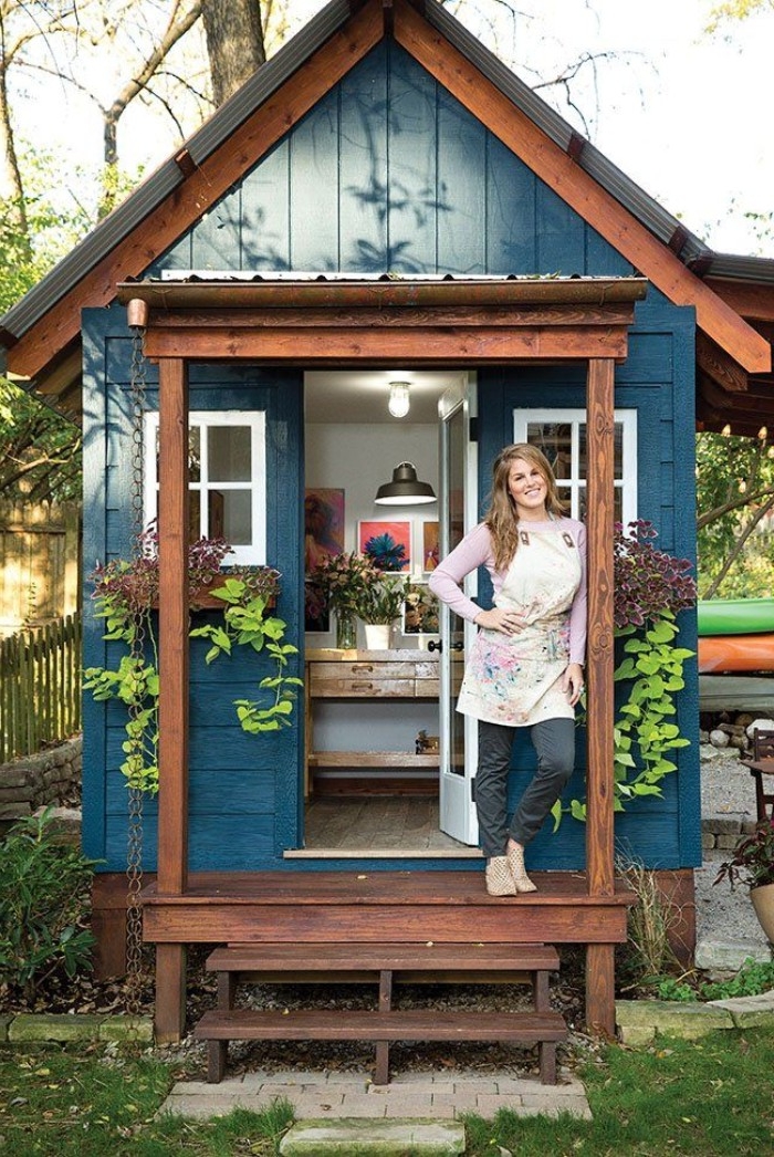 smiling blonde woman, with hand on her hip, standing in front of a blue wooden shed, its door is open, revealing an artist studio inside, she shed interiors, framed artworks and flowers