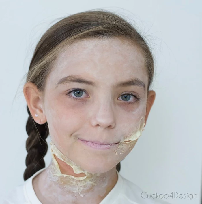 blue-eyed child, with brunette hair, braided in two pigtails, and face covered in powder-like white paint, scary face paint, with fake scars