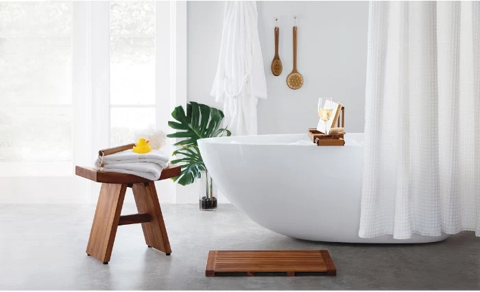 stool made of wood, with two towels, a body brush, and a rubber ducky, placed on a light grey floor, near a smooth white bathtub, bathroom picture ideas, green potted plant, and white curtains
