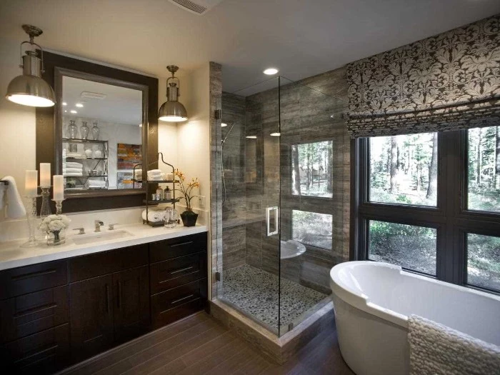 metallic lamps and a square wall mirror, above a dark brown cupboard, with a sink on top, glass shower cabin, and an oval tub nearby