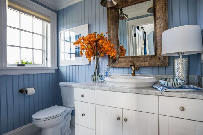 ornamental wooden frame, around a rectangular mirror, mounted on a pale blue wall, with a striped pattern, small bathroom paint colors, white cupboard with a marble countertop, toilet and a window