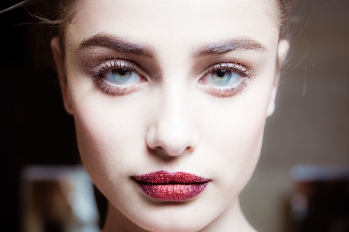 illuminated face of a young pale woman, with light blue eyes, wearing red lipstick, with red glitter