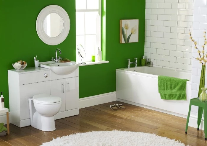 fluffy white rug, on a brown laminate floor, in a bathroom with one visible wall painted green, and the other covered in white subway tiles, white bathtub sink and toilet