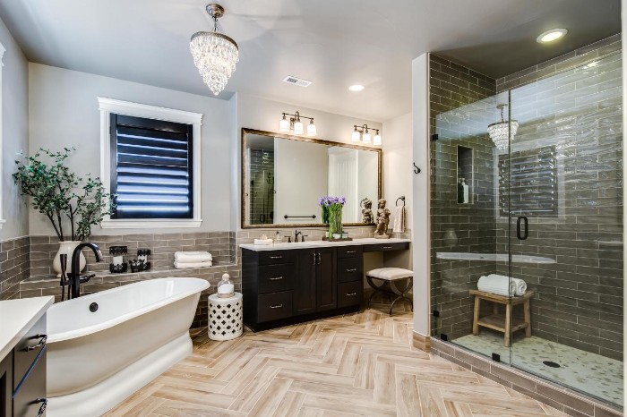 laminate floor in pale beige, inside a bathroom, with dark grey subway tiles, a glass shower cabin, and a vintage-style white bathtub