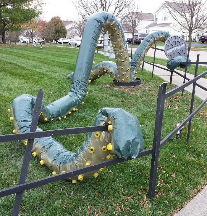 monster with long tentacles, made from inflatable material, climbing out from a fake manhole, scary outdoor halloween decorations, on a large green lawn