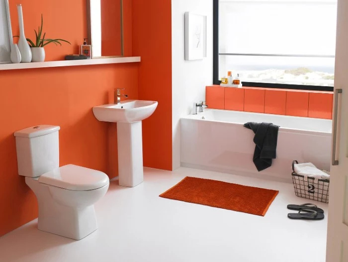 orange bathroom accent wall, in a bright room, containing a large window, a bathtub and a sink, a toilet and a small orange bath mat