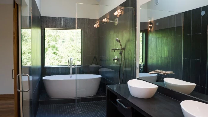 square window overlooking some greenery, inside a room with dark grey tiles, bath remodel ideas, white oval tub, and two white, boat-shaped sinks