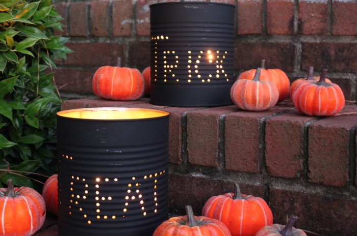 cans painted in black, wit holes spelling the words trick and treat, containing lit candles, haunted house decorations, placed on a brick staircase, with several orange pumpkin ornaments nearby