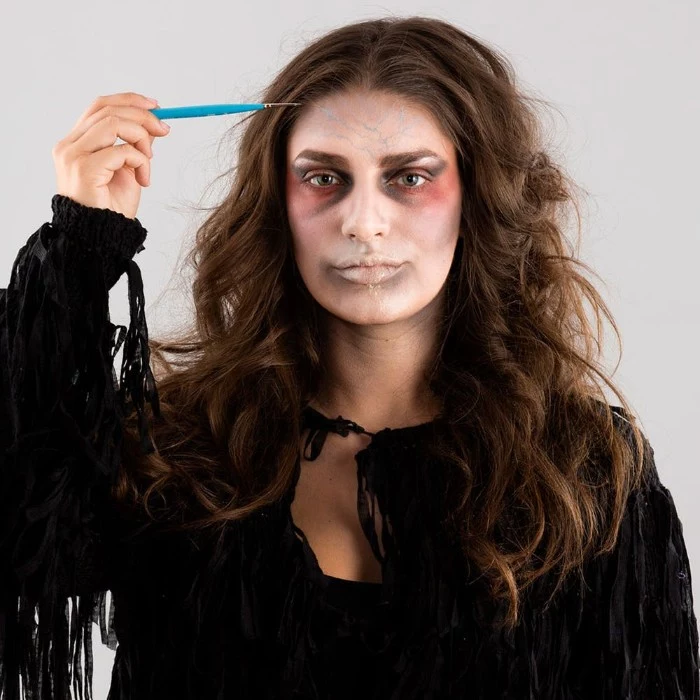 small and thin brush, used to paint little blue and red veins, on the forehead of a woman, wearing zombie face paint, halloween ideas for grownups 