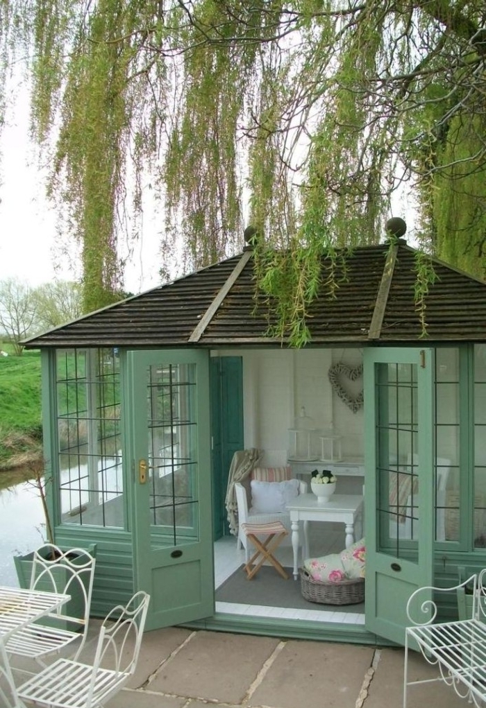 mint green garden shed, with large windows, and open double doors, containing a small white coffee table, a chair and other furniture, weeping willow growing next to it