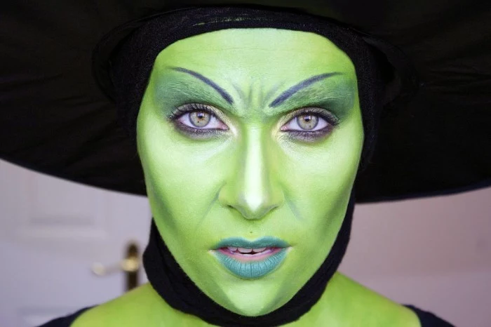 witch face paint, woman with face covered in green paint, wearing bluish green lipstuck, and a large black hat