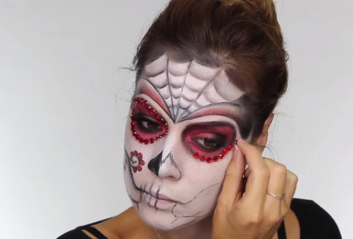 sticking red rhinestones, around the eyes of a young woman, wearing white and black, and pink sugar skull makeup, skeleton face paint, with floral motifs