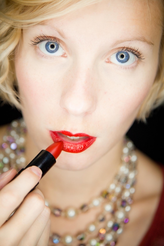 multicolored beaded necklaces, worn by a pale blonde woman, applying red lipstick, while looking at the camera