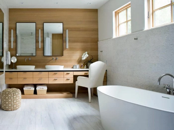 a set of two mirrors, mounted on a wall, covered with wooden panels, oval bathtub in white, cupboards and two swquare windows