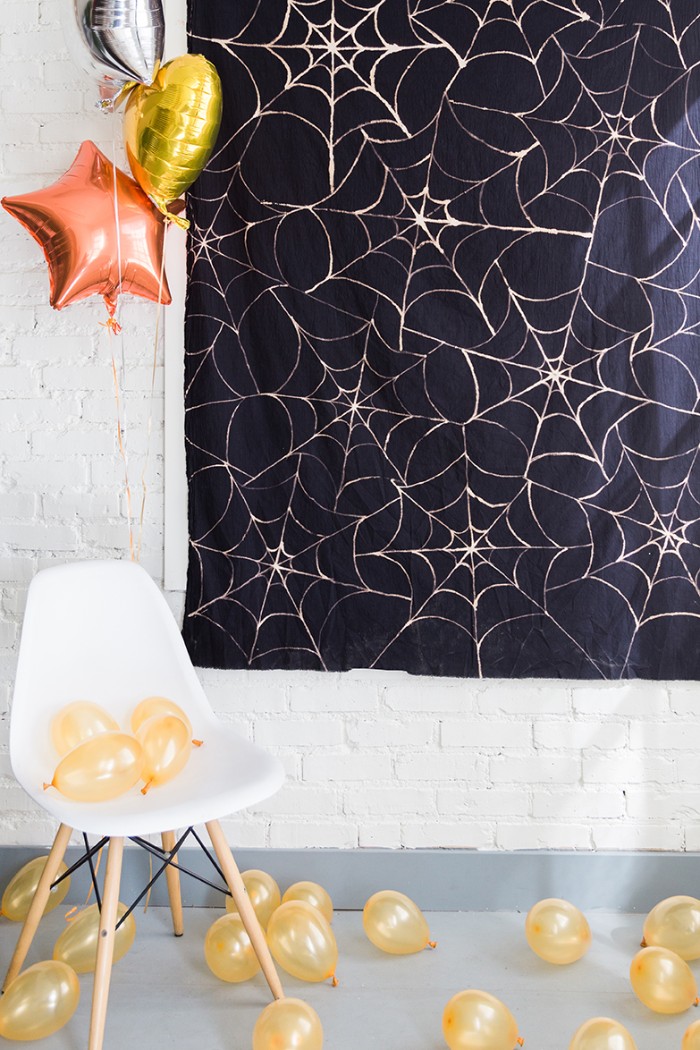 rectangular piece of black fabric, decorated with a pale cobweb pattern, hanging on a white brick wall, halloween decorations, pale yellow ballons, and a white chair, larger balloons shaped like stars and a heart