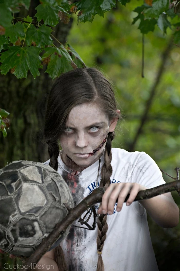 child with long brown pigtails, wearing scary face paint, resembling a zombie, and holding an old football